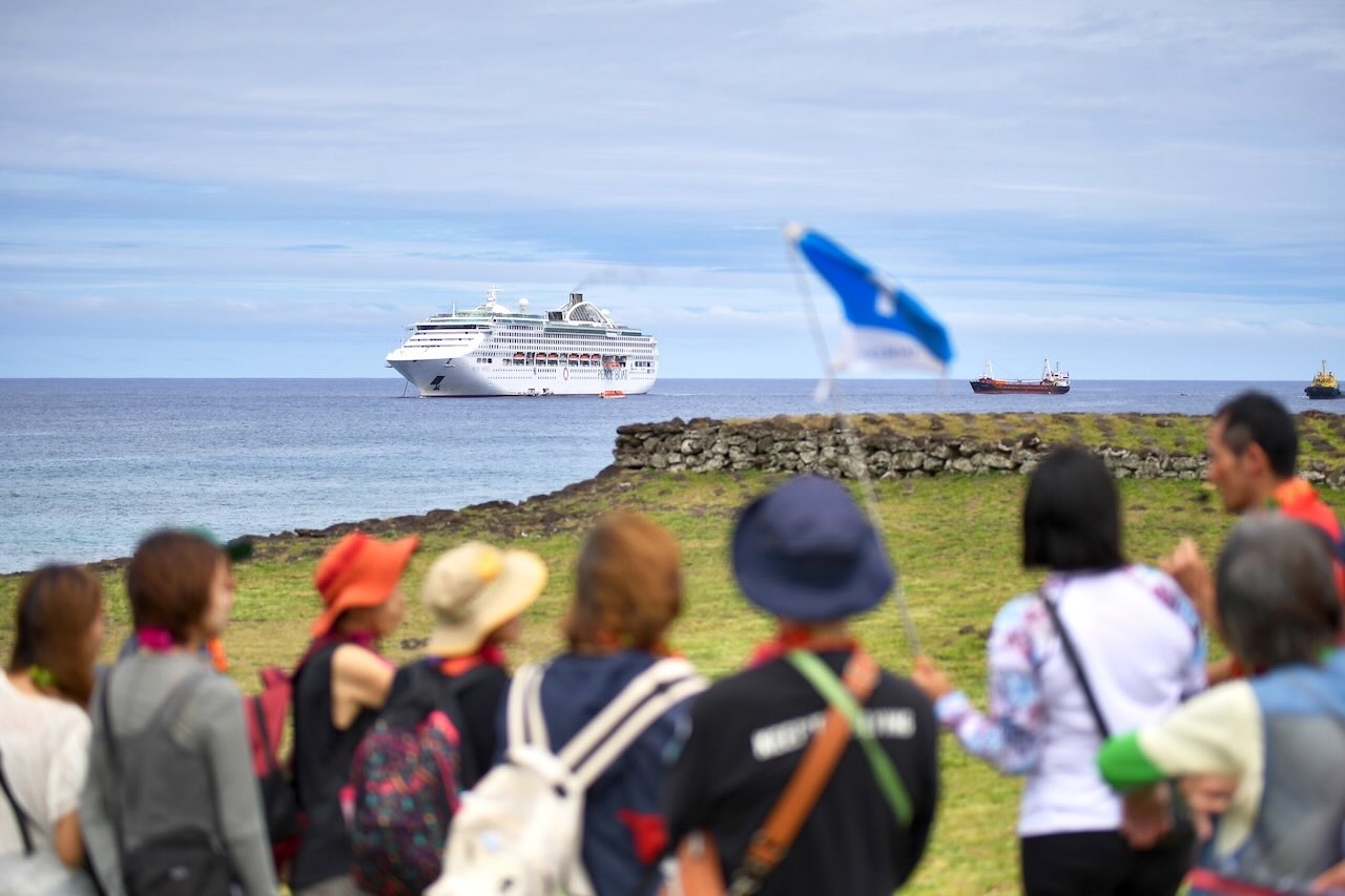 イースター島の草原から沖合に停泊するピースボート「パシフィック・ワールド号」を眺めるツアー参加者たち。ガイドの旗を先頭に、上陸した喜びと一体感が漂う場面。