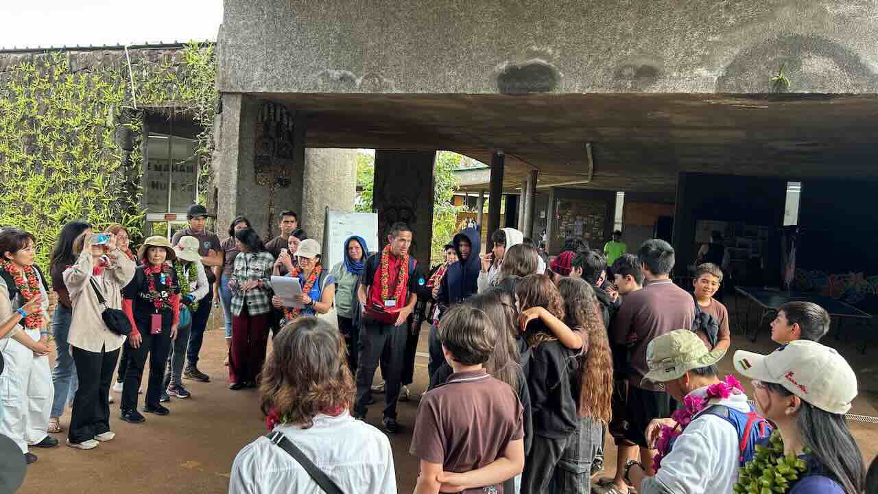 ピースボートの乗客がイースター島の高校を訪れ、現地の生徒たちに支援品を贈呈している様子。レイをかけた参加者と生徒たちの笑顔が交差する、温かい文化交流の瞬間。