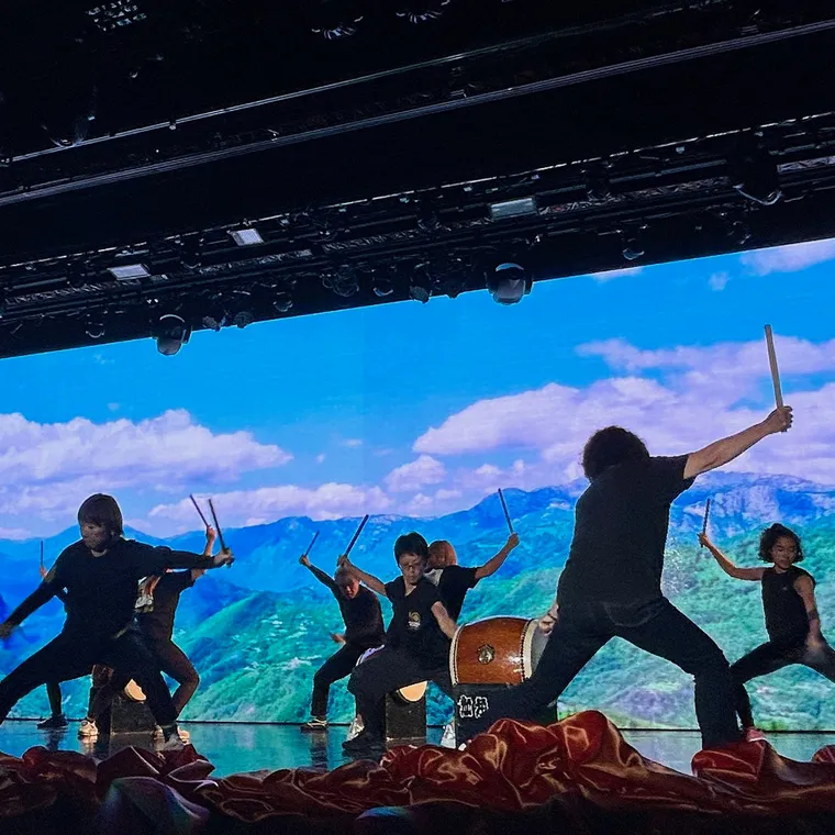 山の映像を背景に舞台で和太鼓を演奏する芸達者祭の出演者たち